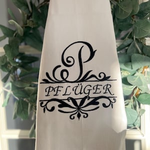 Könnte beinhalten: Weißes Küchentuch mit schwarzem, gesticktem Text und dekorativen Verzierungen. Das Handtuch zeigt den Buchstaben "P" über dem Namen "PFLÜGER" in einer stilisierten Schriftart. Das Handtuch hängt vor einem grünen Kranz.