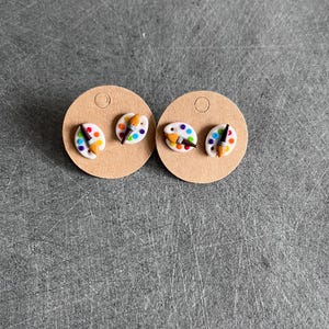 Peut inclure: Une paire de boucles d'oreilles artistiques. Chaque boucle d'oreille présente une mini palette blanche avec des éclaboussures de peinture colorées et un petit pinceau. Les boucles d'oreilles sont présentées sur de petites cartes à boucles d'oreilles rondes et beiges.