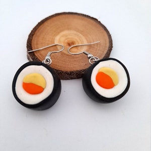 Peut inclure: Une paire de boucles d'oreilles en forme de sushi avec des détails noirs, blancs, orange et jaunes. Les boucles d'oreilles sont en pâte polymère et ont des crochets argentés.