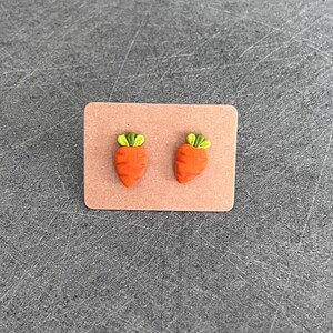 Puede incluir: Un par de pendientes de botón con forma de zanahoria naranja con copas verdes, presentados en una pequeña tarjeta rectangular marrón claro. Los pendientes son un accesorio caprichoso, perfecto para cualquier atuendo.