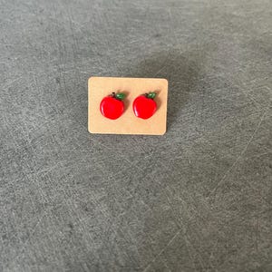 Peut inclure: Une paire de boucles d'oreilles clous en forme de pomme rouge avec des feuilles vertes sur une carte marron.