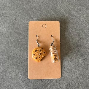 Peut inclure: Une paire de boucles d'oreilles pendantes avec un cookie aux pépites de chocolat et un petit bocal en verre rempli de pépites de chocolat. Les boucles d'oreilles sont présentées sur une carte marron.