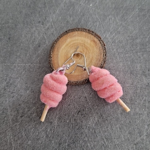 Peut inclure: Boucles d'oreilles en forme de barbe à papa rose avec des bâtonnets en bois et des crochets argentés. Les boucles d'oreilles sont présentées sur une tranche de bois.