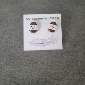 May include: A pair of brown and white ceramic earrings shaped like bottles with a smiling face. The earrings are on a white card with the text "Les Fantaisies d'ATM".