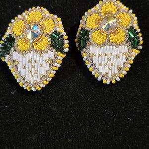 Pode incluir: Um par de brincos de contas com um design de girassol. Os brincos têm pétalas amarelas, uma gema central iridescente e detalhes em branco e dourado. Contas verdes e brancas formam a base.