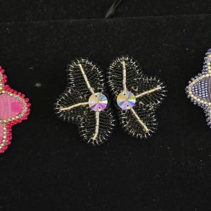 Pode incluir: Três pares de brincos de flores com contas em rosa, preto e azul. Os brincos rosa têm uma borda dourada e uma gema iridescente central. Os brincos pretos têm uma borda prateada e uma gema iridescente central. Os brincos azuis têm uma borda prateada e uma gema iridescente central.