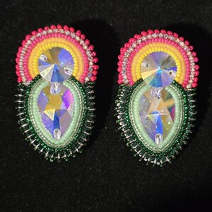 Pode incluir: Um par de brincos de contas com um grande cristal iridescente no centro. Os brincos apresentam um design de contas rosa, amarelo e verde. Os brincos estão sobre um fundo preto.