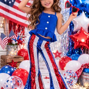 May include: A young person wearing a patriotic outfit. The outfit includes a blue top with white buttons and ruffled hem, and sequined red, white, and blue striped pants. Red heart-shaped sunglasses and a red bow complete the look.