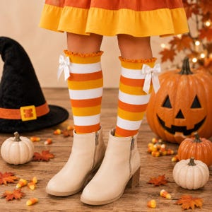 May include: Knee-high socks featuring horizontal stripes in white, orange, and yellow with a ruffled top edge and a small white ribbon bow on the side of each sock. The socks are worn with light beige ankle boots with side zippers.