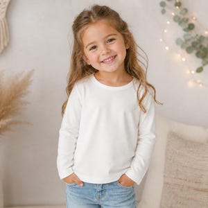May include: A young girl wearing a long-sleeved, off-white shirt and blue jeans smiles at the camera. Her hands are in her pockets. The background includes a decorative wall hanging, dried plants in a vase, and a pillow.