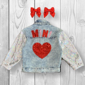 May include: A light blue denim jacket with sequined sleeves and a red heart on the back. The jacket has the word "MINI" in pink and red letters on the back.
