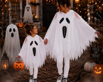 May include: Two white hooded ghost ponchos featuring black oval eyes and a long black mouth graphic. The ponchos have jagged, uneven hemlines and are worn over white pants. One person holds an orange plastic pumpkin bucket with a jack-o'-lantern face.