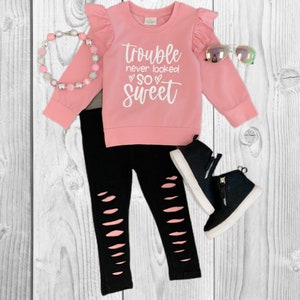 May include: A children's outfit featuring a pink long-sleeved top with ruffle sleeves and the text "Trouble never looked SO SWEET". The outfit includes black ripped leggings, black high-top sneakers, a pink beaded necklace, and iridescent sunglasses.