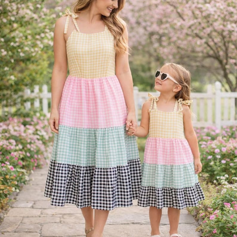 May include: A mother and daughter wearing matching tiered gingham dresses. The dresses feature yellow, pink, mint green, and black and white checkered patterns. The dresses have tie straps and are knee-length. The little girl is wearing heart-shaped sunglasses.