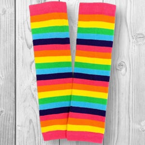 May include: A pair of colorful leg warmers with horizontal stripes in rainbow colors: red, orange, yellow, green, blue, and purple. The top and bottom cuffs are bright pink. The leg warmers are displayed against a white wooden background.