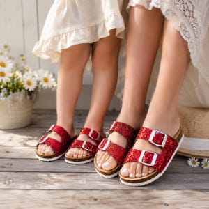 May include: Red glitter sandals with two adjustable straps and silver buckles. The sandals have a cork footbed and white soles. The image shows a pair of adult sandals and a pair of children's sandals.