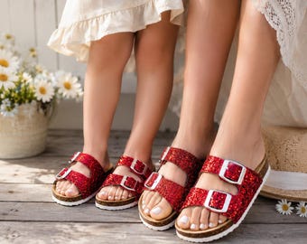 May include: Red glitter sandals with two adjustable straps and silver buckles. The sandals have a cork footbed and white soles. The image shows a pair of adult sandals and a pair of children's sandals.
