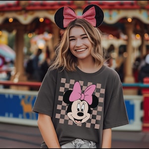 Op de afbeelding: Een vrouw draagt een grijs T-shirt met een geruit patroon en een roze Minnie Mouse-afbeelding. Ze draagt een rood-wit geblokt hoofdband met muisoren.