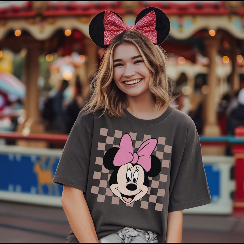 Op de afbeelding: Een vrouw draagt een grijs T-shirt met een geruit patroon en een roze Minnie Mouse-afbeelding. Ze draagt een rood-wit geblokt hoofdband met muisoren.