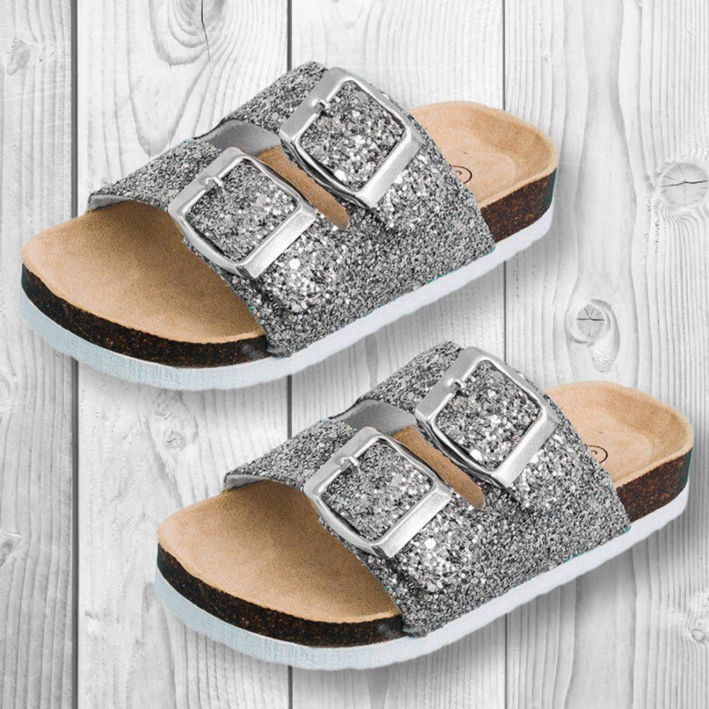 May include: Two pairs of silver glitter sandals with adjustable silver buckles. The sandals have a cork footbed and a white sole. The sandals are designed for casual wear and are suitable for warm weather.