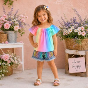 May include: A rainbow-colored short-sleeve top with a scoop neckline and a matching hair bow, paired with denim shorts featuring a vertical rainbow sequin design and a frayed hem, and light-colored sandals with two straps and metal buckles.