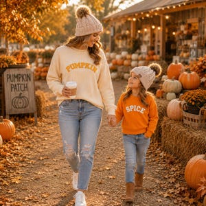 May include: A cream-colored long-sleeve sweater with the word PUMPKIN in orange, paired with light blue distressed jeans and a matching knit beanie. Beside it, an orange long-sleeve sweater with the word SPICE, light blue jeans, and a knit beanie.