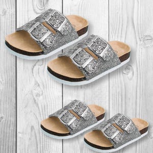 May include: Four pairs of silver glitter sandals with adjustable silver buckles. The sandals have a cork footbed and a black sole. The sandals are displayed on a white wooden background.
