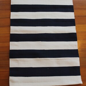 May include: A navy blue and white striped rug with a woven texture. The rug has a simple design and is perfect for adding a touch of nautical style to any room.