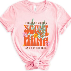 K&ouml;nnte beinhalten: Rosafarbenes T-Shirt mit dem Text "Fueled by Cookies Scout Mama and Adventures" in orangefarbener und brauner Schrift mit gr&uuml;nen Bl&auml;ttern und rosa Bl&uuml;ten.
