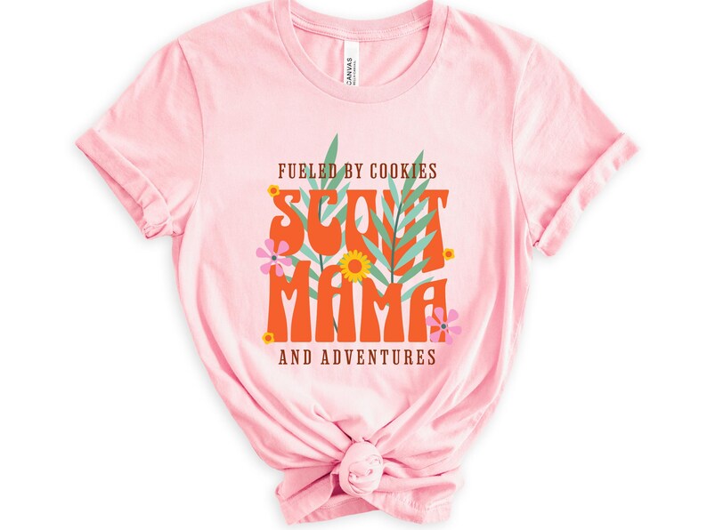 K&ouml;nnte beinhalten: Rosafarbenes T-Shirt mit dem Text "Fueled by Cookies Scout Mama and Adventures" in orangefarbener und brauner Schrift mit gr&uuml;nen Bl&auml;ttern und rosa Bl&uuml;ten.