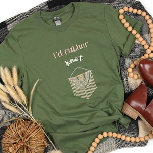 May include: An olive green t-shirt with the text "I'd rather Knot" and a macrame design. The shirt is styled with brown shoes, a wooden bead garland, and a black and grey plaid blanket.