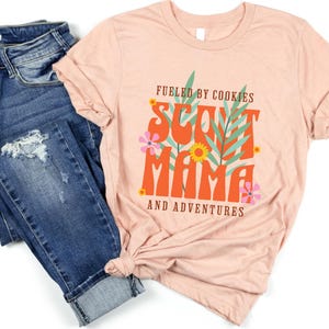 K&ouml;nnte beinhalten: Ein pinkfarbenes T-Shirt mit dem Text "Fueled by Cookies Scout Mama and Adventures" in orangefarbener und brauner Schrift mit floralen Akzenten.
