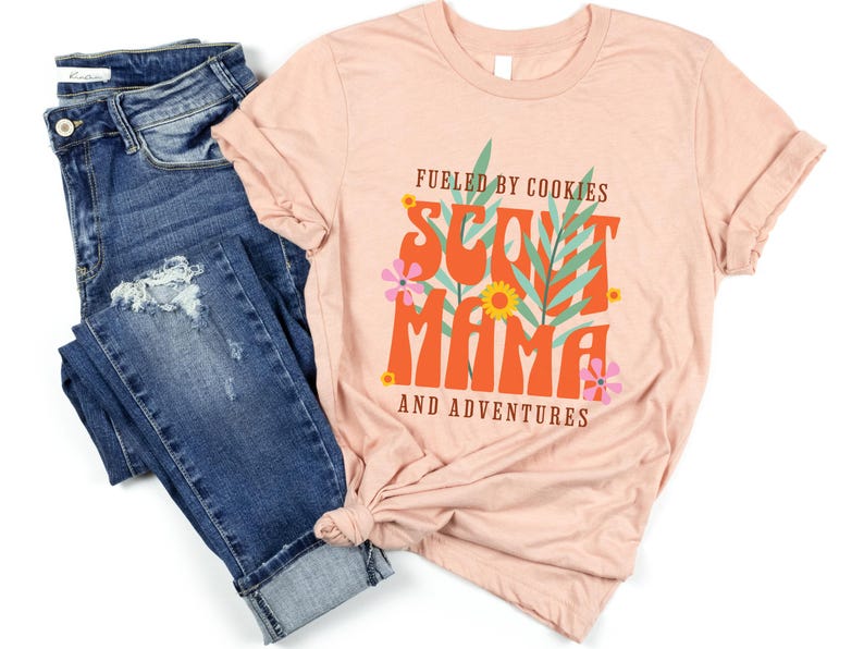 K&ouml;nnte beinhalten: Ein pinkfarbenes T-Shirt mit dem Text "Fueled by Cookies Scout Mama and Adventures" in orangefarbener und brauner Schrift mit floralen Akzenten.