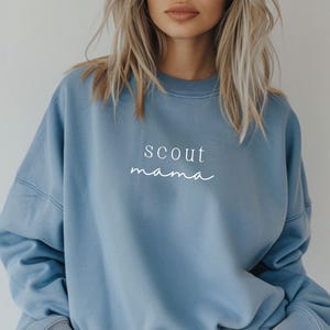 Puede incluir: Una sudadera de color azul claro con el texto "scout mama" impreso en blanco en la parte delantera.
