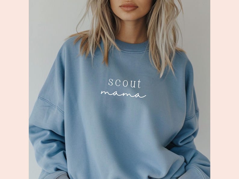 Puede incluir: Una sudadera de color azul claro con el texto "scout mama" impreso en blanco en la parte delantera.