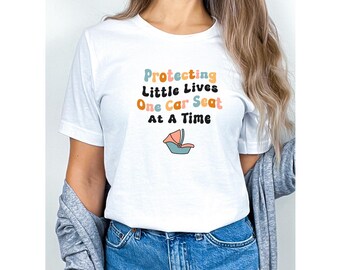 May include: White t-shirt with the text "Protecting Little Lives One Car Seat At A Time" in colorful, rounded letters. A cartoon car seat is pictured below the text.
