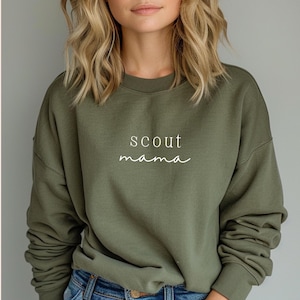 Puede incluir: Una sudadera verde con el texto "scout mama" en letras blancas.