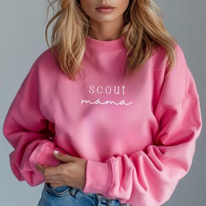 Puede incluir: Sudadera rosa con el texto "scout mama" en letras blancas.