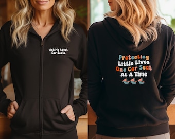 Könnte beinhalten: Zwei schwarze Kapuzenpullover mit Reißverschluss und unterschiedlichen Slogans. Der erste Hoodie sagt "Ask Me About Car Seats". Der zweite Hoodie sagt "Protecting Little Lives One Car Seat At A Time" mit drei bunten Autositz-Symbolen.