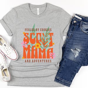 K&ouml;nnte beinhalten: Graues T-Shirt mit dem Text "Fueled by Cookies Scout Mama and Adventures" in Orange und Gr&uuml;n mit floralen Akzenten.