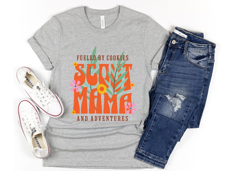 K&ouml;nnte beinhalten: Graues T-Shirt mit dem Text "Fueled by Cookies Scout Mama and Adventures" in Orange und Gr&uuml;n mit floralen Akzenten.