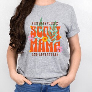 K&ouml;nnte beinhalten: Graues T-Shirt mit einem grafischen Design, auf dem "Fueled by Cookies Scout Mama and Adventures" steht. Die Grafik ist in den Farben Orange, Gr&uuml;n und Pink gehalten.