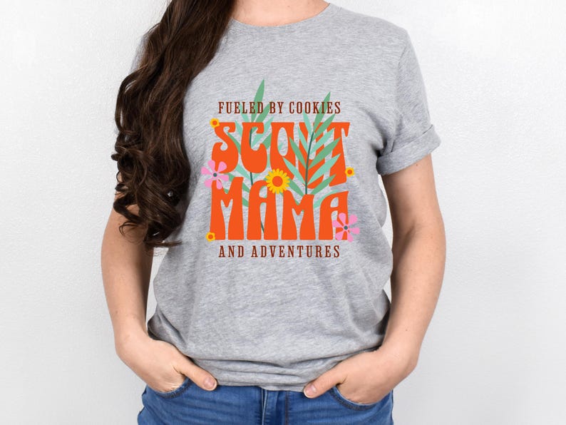 K&ouml;nnte beinhalten: Graues T-Shirt mit einem grafischen Design, auf dem "Fueled by Cookies Scout Mama and Adventures" steht. Die Grafik ist in den Farben Orange, Gr&uuml;n und Pink gehalten.