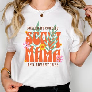 K&ouml;nnte beinhalten: Wei&szlig;es T-Shirt mit dem Text "Fueled by cookies Scout Mama and adventures" in orangefarbener und gr&uuml;ner Schrift mit floralen Akzenten.