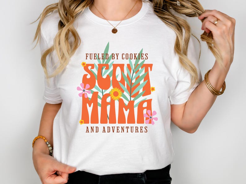 K&ouml;nnte beinhalten: Wei&szlig;es T-Shirt mit dem Text "Fueled by cookies Scout Mama and adventures" in orangefarbener und gr&uuml;ner Schrift mit floralen Akzenten.