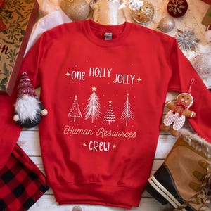 Peut inclure: Un sweat-shirt rouge avec l'inscription blanche "one HOLLY JOLLY Human Resources CREW". Le motif comprend des sapins et des étoiles blancs. Le sweat-shirt est entouré de décorations de Noël, dont un gnome, un bonhomme en pain d'épices et des bottes.