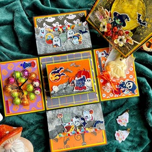 May include: A multi-panel Halloween-themed scrapbook with various decorative elements. The panels feature illustrations of ghosts, pumpkins, bats, and tombstones. The color palette includes orange, black, gray, and purple. The scrapbook is open, revealing several decorated pages.