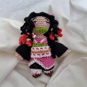 May include: A handmade crochet doll featuring a character with black hair, pink eyes, and a green mouth. The doll wears a pink and white kimono with a black jacket. Red accents are present in the hair and clothing. The doll is approximately 5 inches tall.