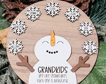 May include: A wooden snowman ornament with the text "GRANDKIDS are like snowflakes each one is beautiful and unique".  The snowman has eight snowflakes around it, each with a name written on it: Graeden, William, Sienna, Olivia, Logan, Kaia, Isla.