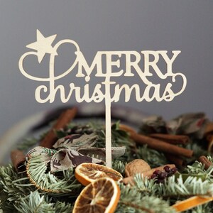 May include: A wooden sign with the words "Merry Christmas" in a cursive font, with a star on top. The sign is placed in a wreath of pine branches, cinnamon sticks, dried oranges, and walnuts.